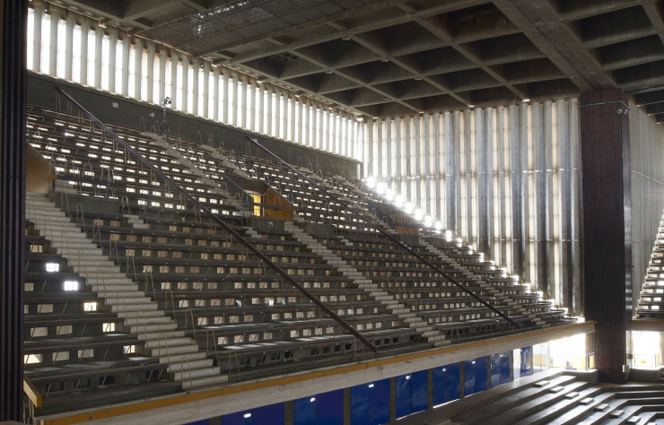 Dans le stade couvert, au sein du complexe sportif, les gradins sont conçus comme des claustras. De l’eau courante refroidit les sièges par en dessous. Des trous percés transforment trois des murs en écrans lumineux, offrant un éclairage doux et une ventilation croisée. Un effet dedans-dehors : la construction écologique avant l’heure.