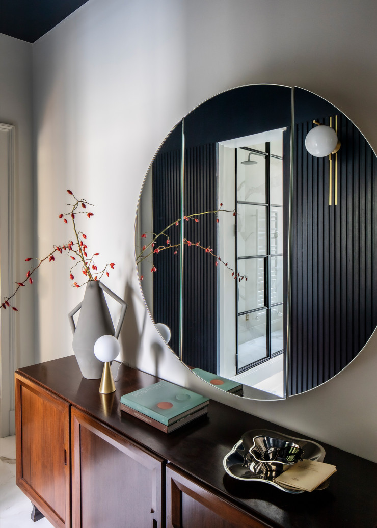 Dans le miroir rond conçu en triptyque par le propriétairee se reflète le lambris en bois fraisé et teinté du couloir. L’applique IC Lights, de Michael Anastassiades (Flos), est fixée sur des insertions verticales en laiton. Sur le buffet vintage, de Franco Albini (Poggi), vase Kora, design Studiopepe (Atipico), lampe Bilia Mini, de Gio Ponti (Fontana Arte), et plats en acier conçus par Alvar Aalto (Iittala). Sol en grès cérame Dreaming (Lea Ceramiche).