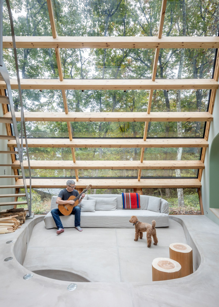 Le salon occupe une place centrale dans la maison. Entièrement vitré, il exprime la volonté forte de faire corps avec la nature, en évitant toute position de surplomb. La structure est réalisée en cèdre du Japon. Le canapé en lin, sur lequel Robert Tull, le propriétaire, est assis, a été dessiné spécifiquement pour le projet par l’architecte. Tabourets en rondins de bois faits maison.