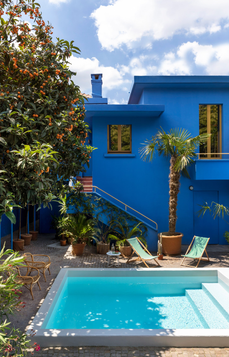 Ceinturée de néfliers et de palmiers, la villa s’articule autour d’une piscine centrale. Sa façade bleu électrique, hommage vibrant à la Casa Azul de Frida Kahlo et au jardin Majorelle de Marrakech, transforme la cour industrielle en un éden subtropical. Fauteuils, transats et table basse vintage.